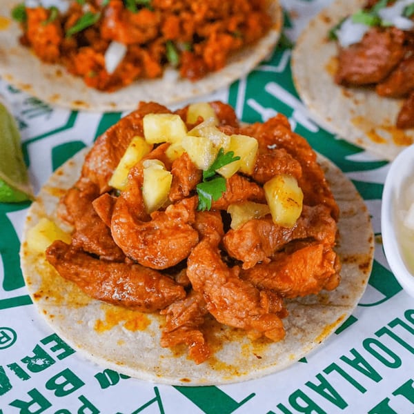 Al pastor taco with marinated pork and pineapple on a tortilla, with other tacos blurred in the background.