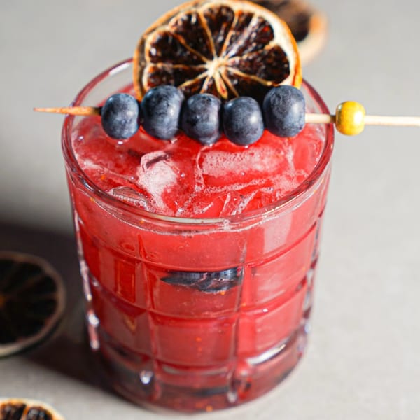 A vibrant red cocktail with ice, garnished with blueberries on a skewer and a dried orange slice.