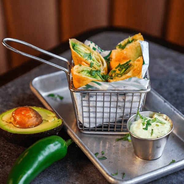Guapo's avocado egg rolls in a metal basket with a side of dipping sauce, jalapeño, and avocado.