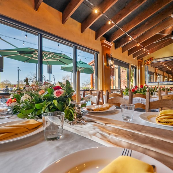 Set tables with floral centerpieces, yellow napkins, and large windows overlooking outdoor patio umbrellas.