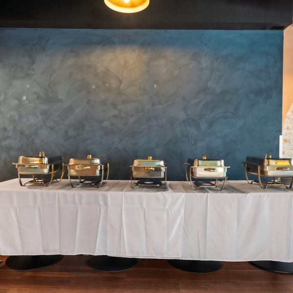 Five chafing dishes on a white-covered buffet table against a textured blue wall.