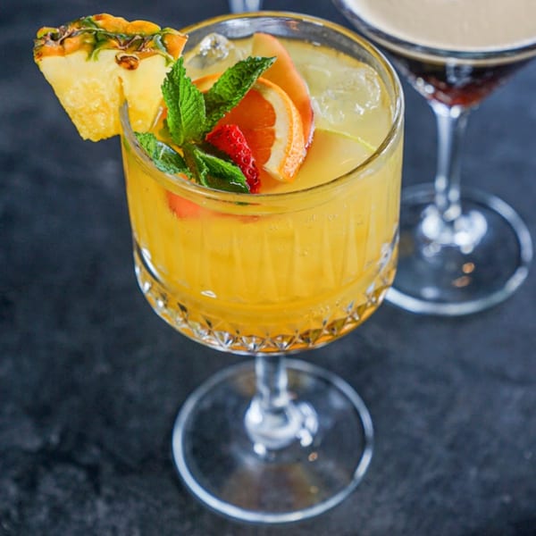 A vibrant yellow cocktail garnished with pineapple, orange slices, strawberry, and mint leaves.