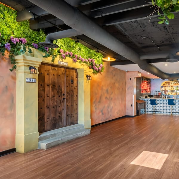 Restaurant interior with a wooden door, green foliage, purple flowers, and a tiled bar area.