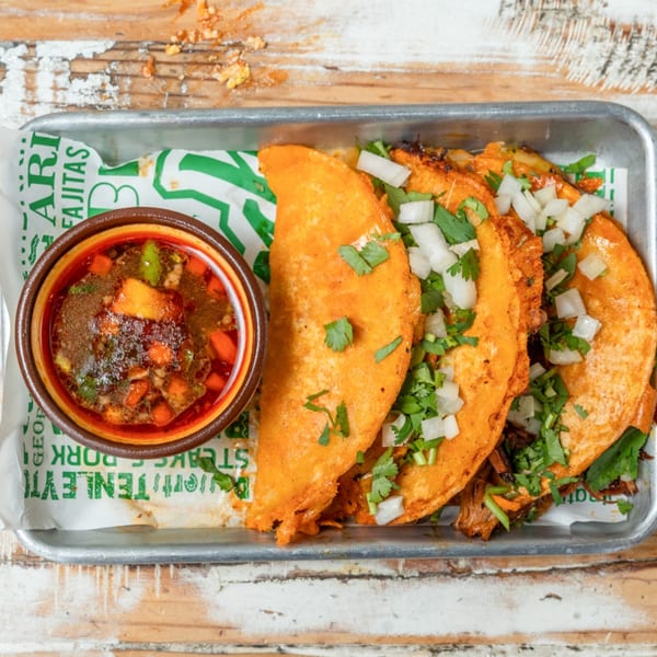 Three birria tacos with cilantro and onions, served with a side of consommé on a metal tray.