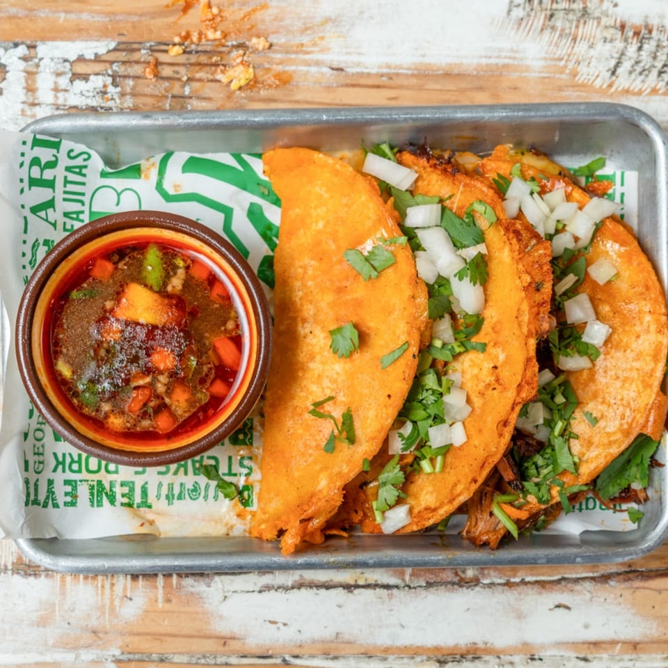 Three birria tacos with cilantro and onions, served with a side of consommé on a metal tray.
