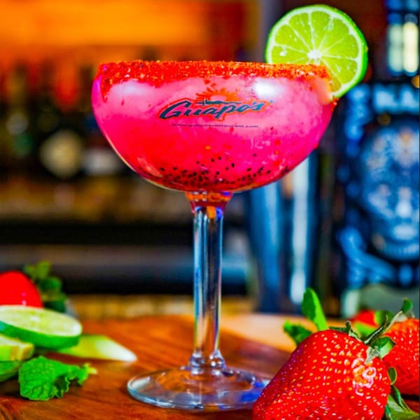 A vibrant pink Guapo's margarita with a chili-lime rim and lime wedge, surrounded by fresh strawberries.