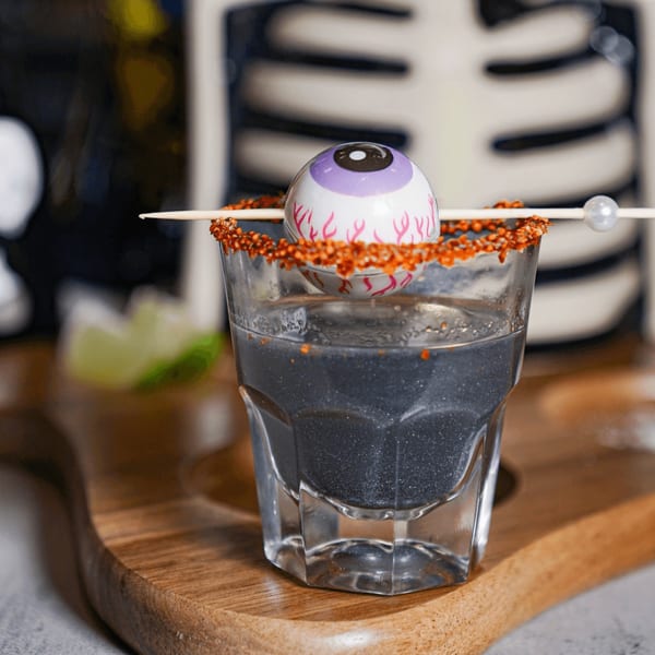 A Halloween-themed black cocktail with a chili-salted rim and an eyeball garnish on a wooden board.
