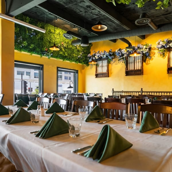 Restaurant interior with yellow walls, green plant decor, dark wood chairs, and tables set with green napkins.