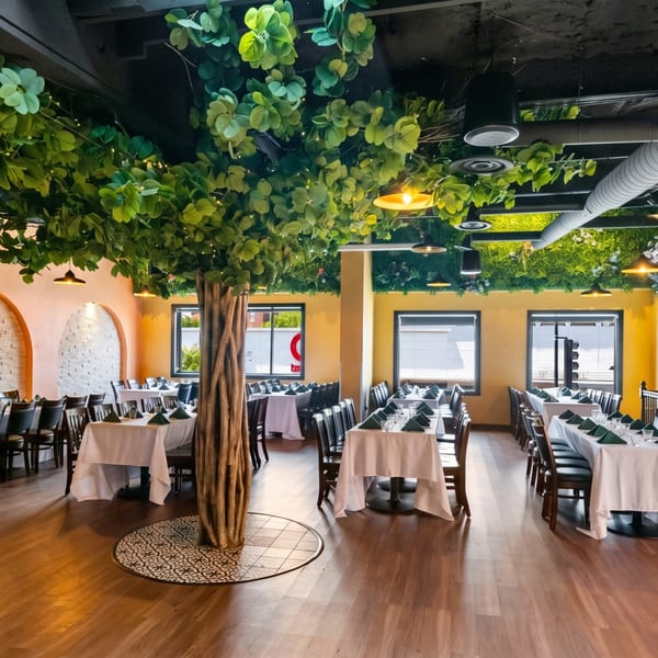 Restaurant dining room with tables set, a large artificial tree, and green foliage on the ceiling.