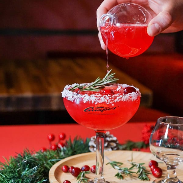 Hand pouring red liquid from a spherical bottle into a Guapo's margarita glass with rosemary and cranberries.
