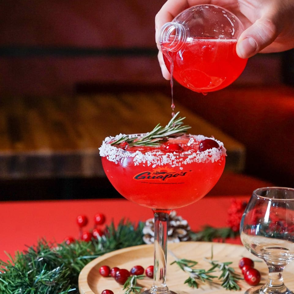 Hand pouring red liquid from a spherical bottle into a Guapo's margarita glass with rosemary and cranberries.