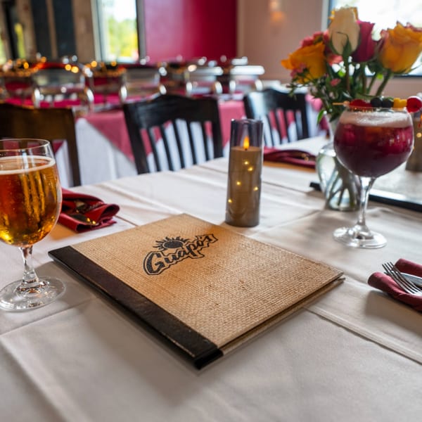 A Guapo's menu, beer, and cocktail on a white tablecloth with a floral centerpiece.