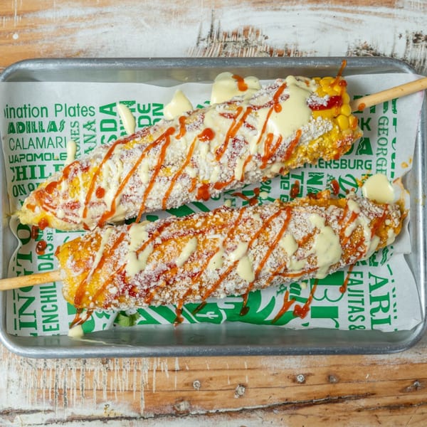 Two Mexican street corn (elotes) on a tray, topped with white sauce, red drizzle, and cheese.